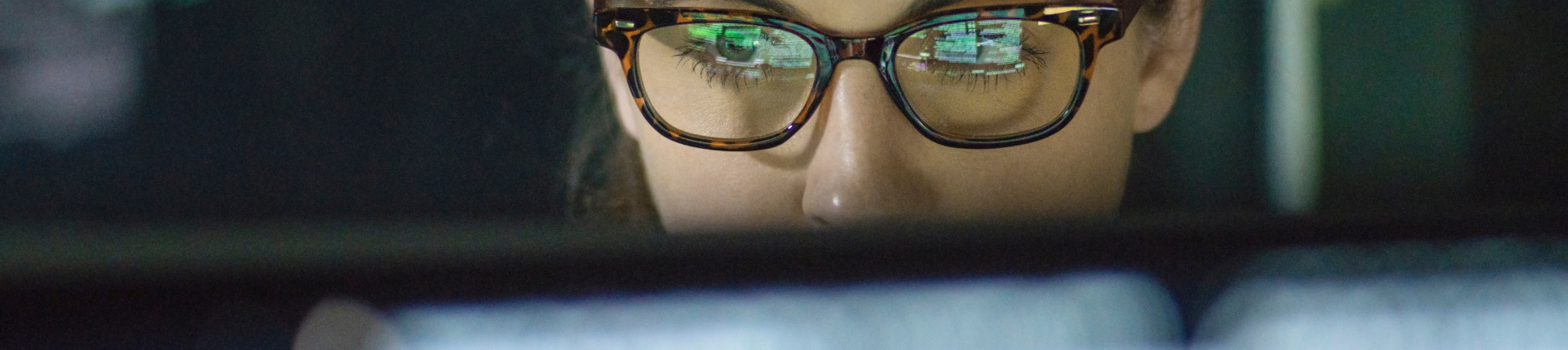 Image shows the top half of a woman wearing glasses looking at her laptop, rest of her face covered by laptop