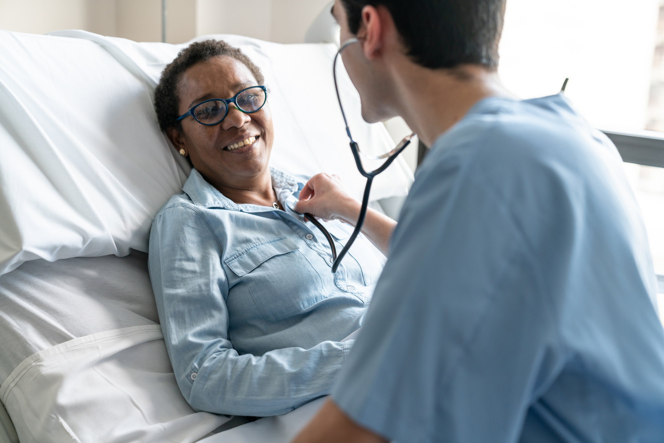 Female black patient lying down on hospital bed while nurse is checking her heart beat with stethoscope